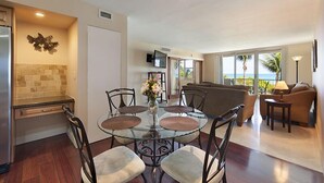A dining area with a glass table, chairs, and a vase of flowers. There is a kitchen with a refrigerator and a tiled backsplash. A living area with a sofa and a lamp is visible. - Spectacular Oceanfront Views in Miami's Safest Beachfront Community  (Surfside)