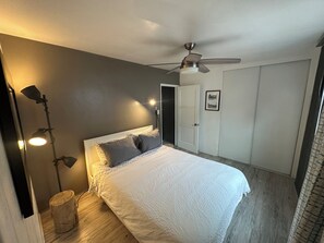 Un dormitorio con cama, ventilador de techo, lámpara de noche, puerta y un pequeño taburete de madera.