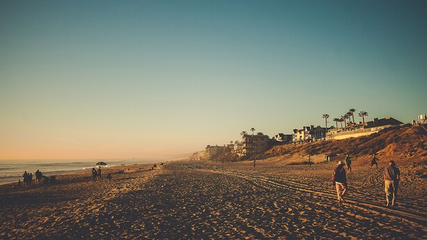 A beach with people walking and sitting, buildings on a hillside, and a clear sky.