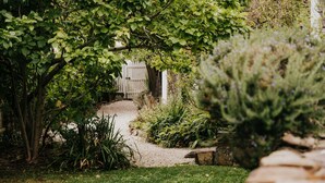 Ein Gartenweg führt zu einem hölzernen Tor, umgeben von üppigem Grün und verschiedenen Pflanzen.