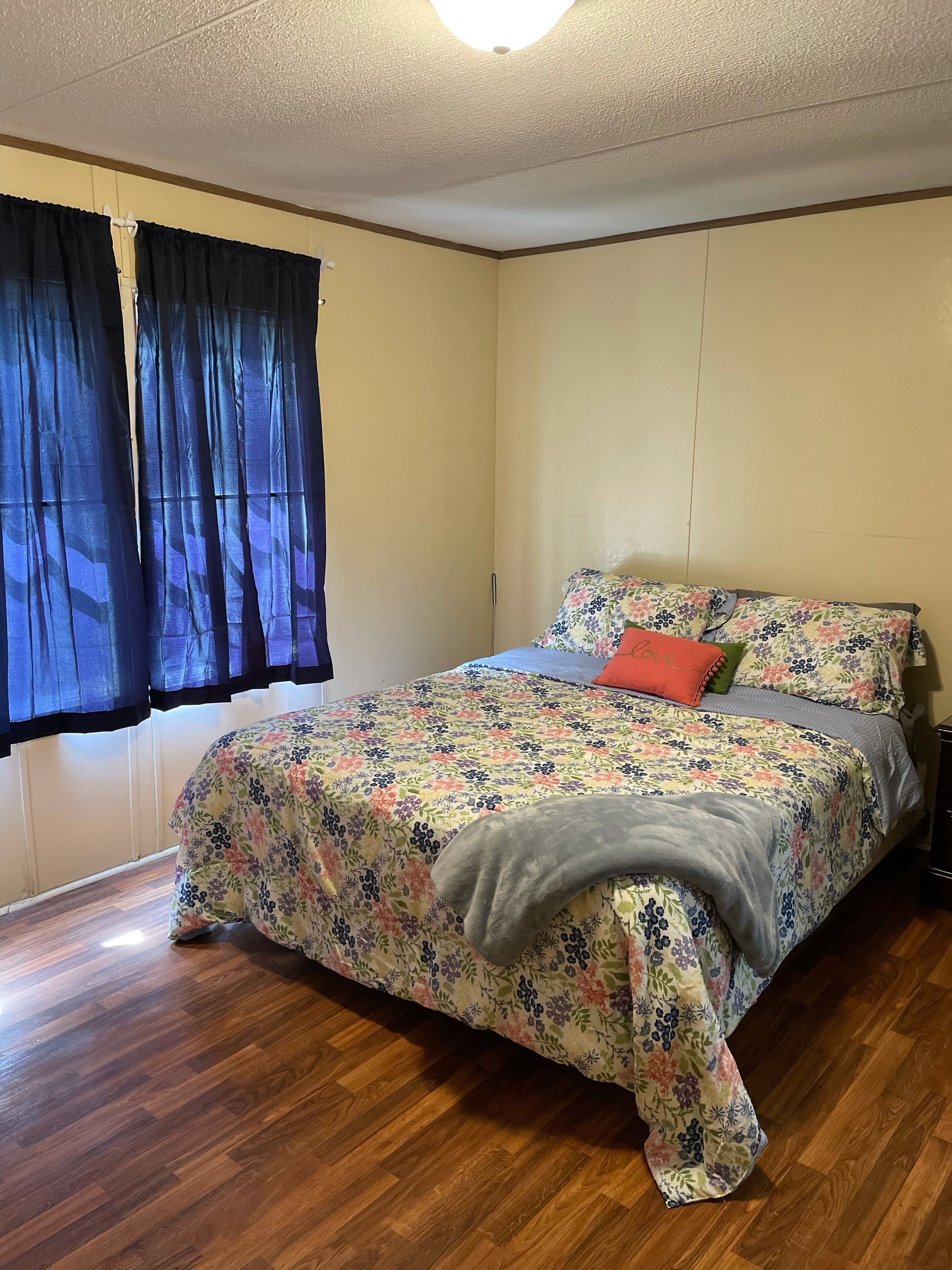 A bedroom with a floral bedspread, a wooden floor, and a window with dark curtains.