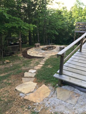 Un chemin de pierres mène à une terrasse en bois où se trouve un foyer au centre.