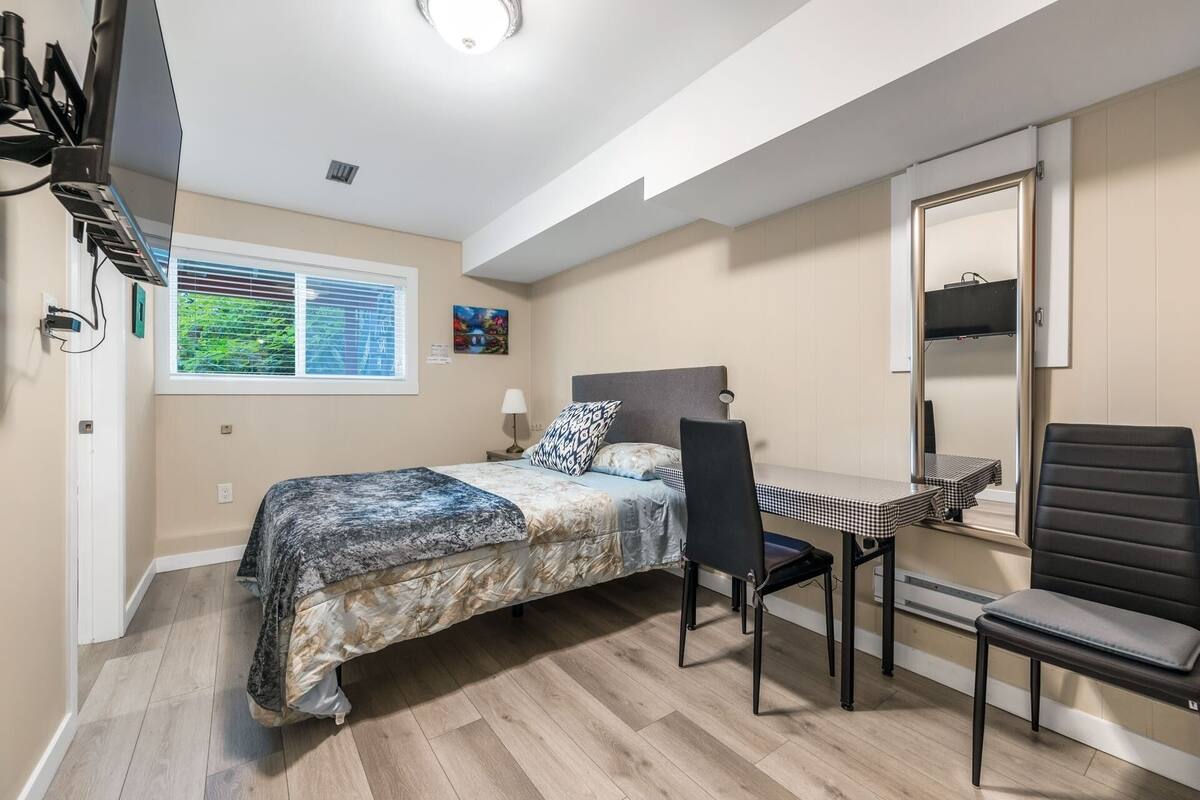 Ein Schlafzimmer mit einem Bett, einem Schreibtisch und einem Stuhl. Es gibt ein Fenster mit Jalousien, eine Lampe und ein Bild an der Wand.