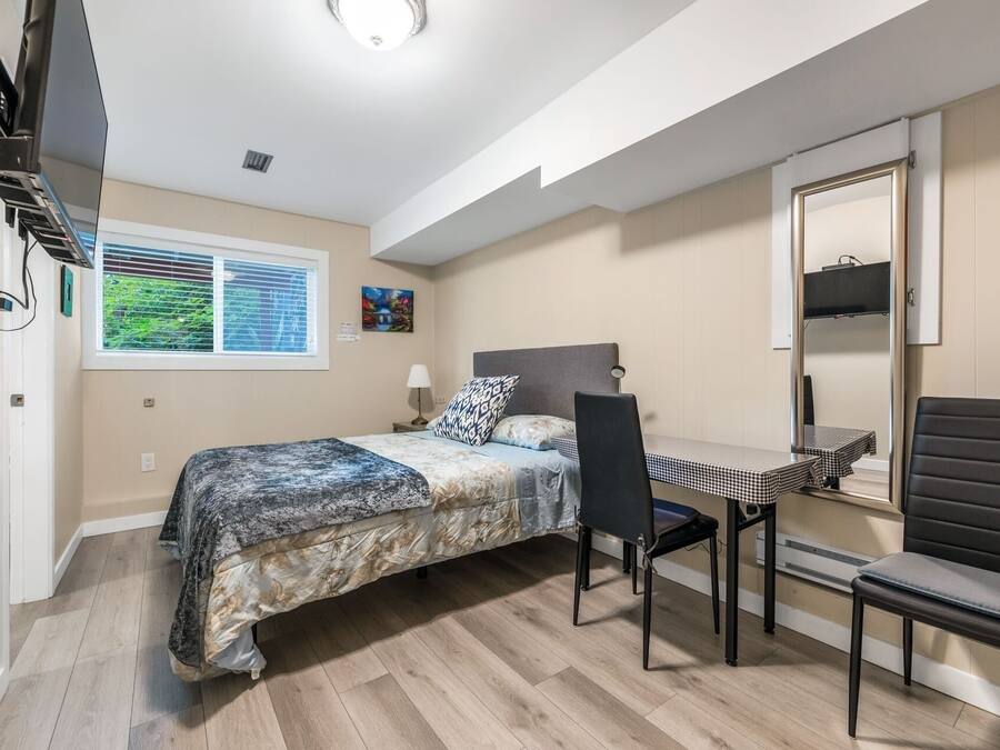 A bedroom with a bed, a desk, and a chair. There is a window with blinds, a lamp, and a painting on the wall.