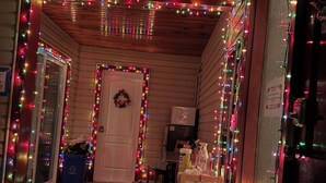 Ein Flur mit Holzwänden und -decke, dekoriert mit bunten Lichterketten. Eine Tür ist mit einem Kranz geschmückt, ein Tisch ist mit Gegenständen bestückt und ein Stuhl steht daneben.
