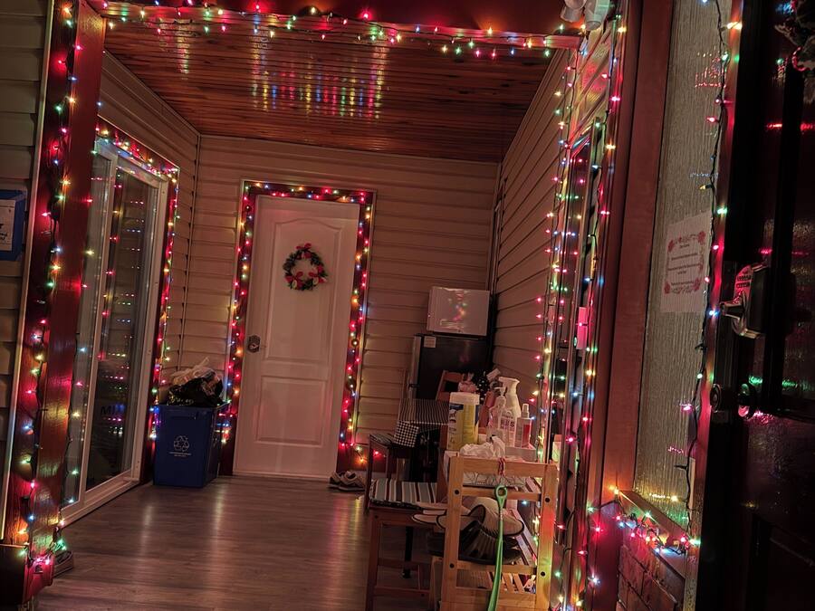 Un pasillo con paredes y techo de madera, decorado con luces de colores. Se observa una puerta adornada con un adorno de ramas, una mesa con objetos y una silla.