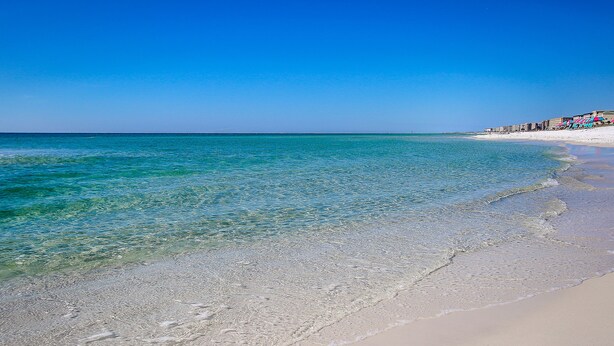 A beach with clear turquoise water, a sandy shore, and a row of buildings with colorful umbrellas. - 🦩Book Me Baby! Gulf-Front Condo With Gorgeous Views 🦩 (Fort Walton Beach)
