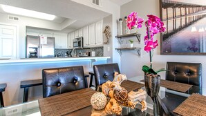 A modern kitchen with white cabinets, a stainless steel refrigerator, and a microwave. There is a dining area with a glass table, brown placemats, and a vase with pink orchids. - 🦩Book Me Baby! Gulf-Front Condo With Gorgeous Views 🦩 (Fort Walton Beach)