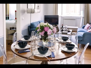 Ein Essbereich mit einem runden Tisch, gedeckt für sechs Personen, umgeben von transparenten Stühlen. Auf dem Tisch steht eine Vase mit Blumen, im Hintergrund ist ein Fernseher zu sehen.