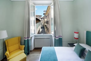 A hotel room with a bed, a yellow armchair, a view of a narrow street through an open window, and a radiator.