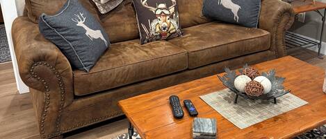 A cozy living room with a brown leather couch, two decorative pillows, a framed landscape painting, a wooden coffee table with remote controls and decorative items, a patterned rug, and a side table with a lamp.