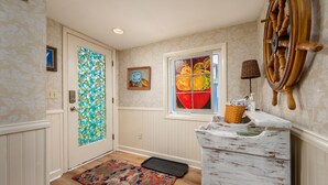 A hallway with a wooden floor, a patterned rug, a door with a stained glass window, a wall-mounted lamp, and a decorative wheel.