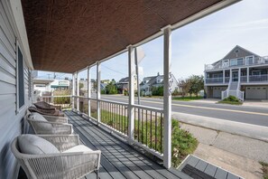 Un porche cubierto con muebles de mimbre, vista a la calle y un edificio de dos pisos con garaje.