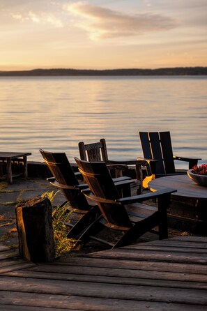 Una terraza de madera con sillas Adirondack y una mesa, con vista a una masa de agua tranquila durante el atardecer.