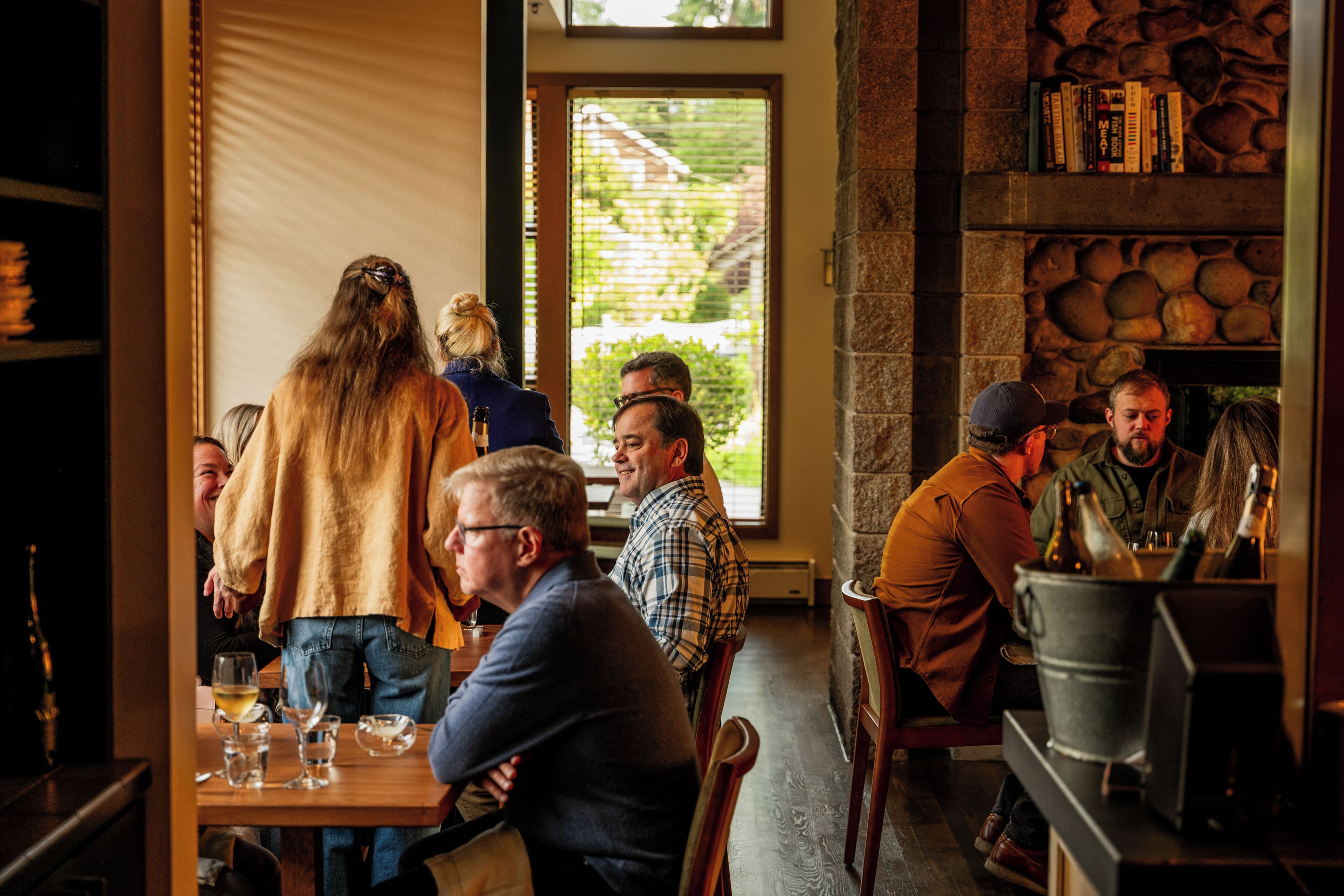 Een groep mensen zit aan een houten tafel in een restaurant, met een stenen muur en een raam op de achtergrond.