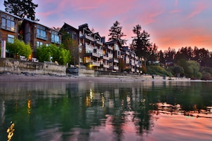 A waterfront hotel with multiple balconies, a reflective water surface, and a backdrop of trees and a sunset sky.