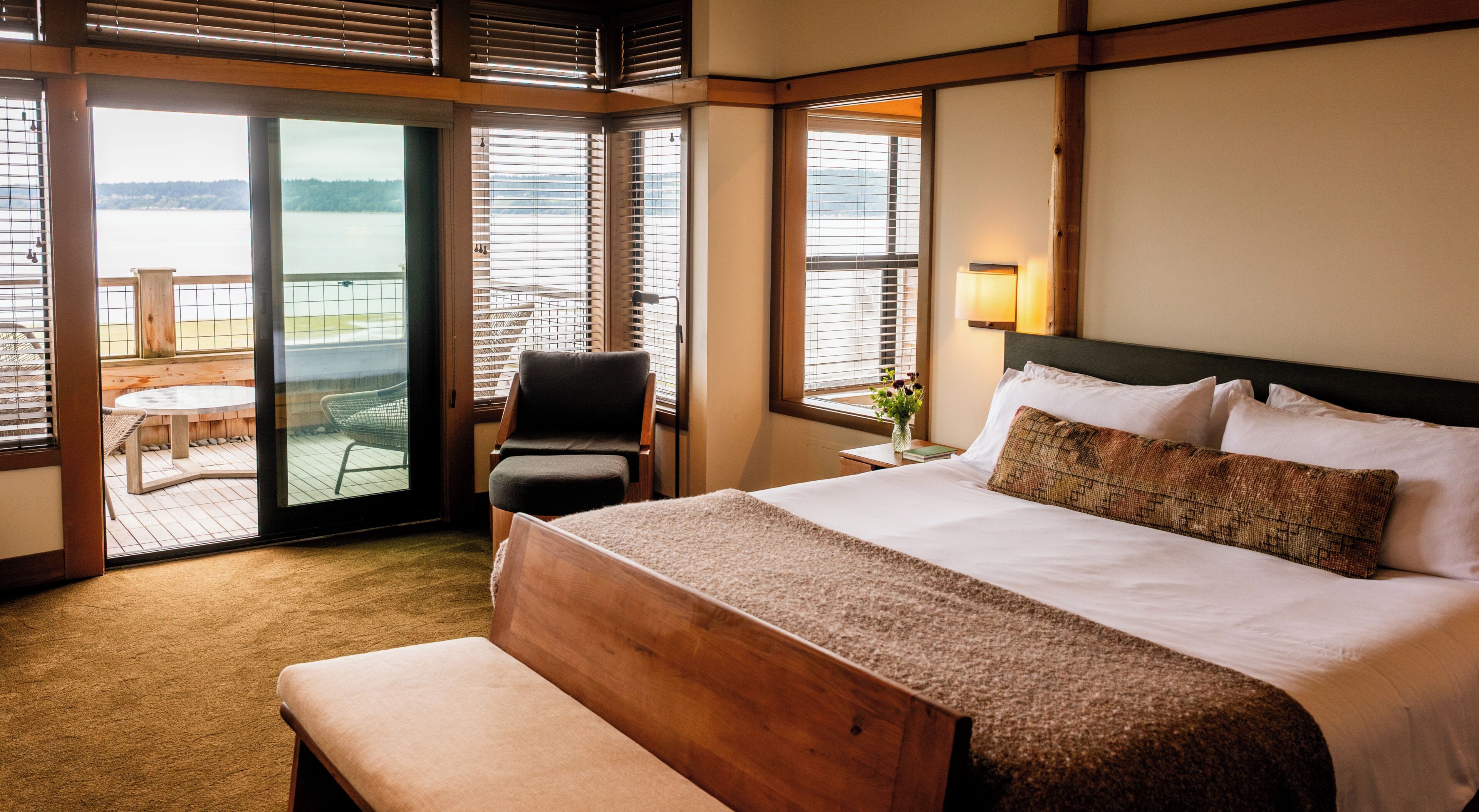 A hotel room with a large bed, a bench, a chair, and a view of a balcony with a table and chairs.