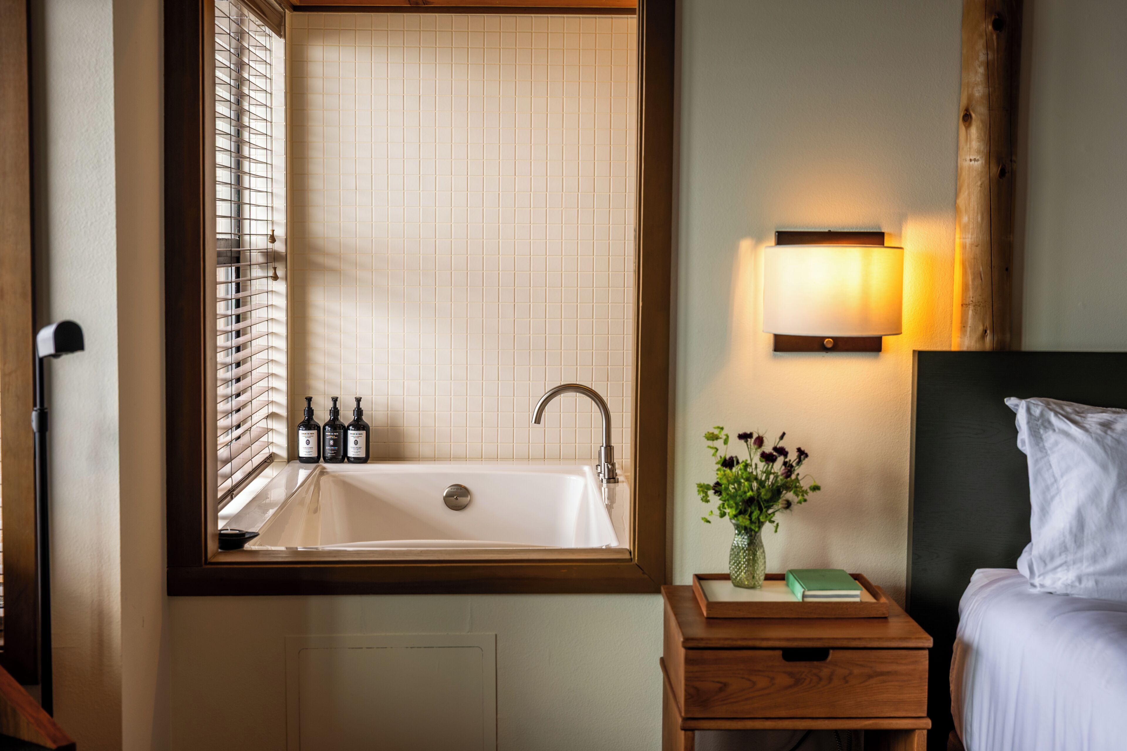 A bathroom with a bathtub, a mirror, a window with blinds, a bedside table with a lamp, and a nightstand with a vase of flowers.