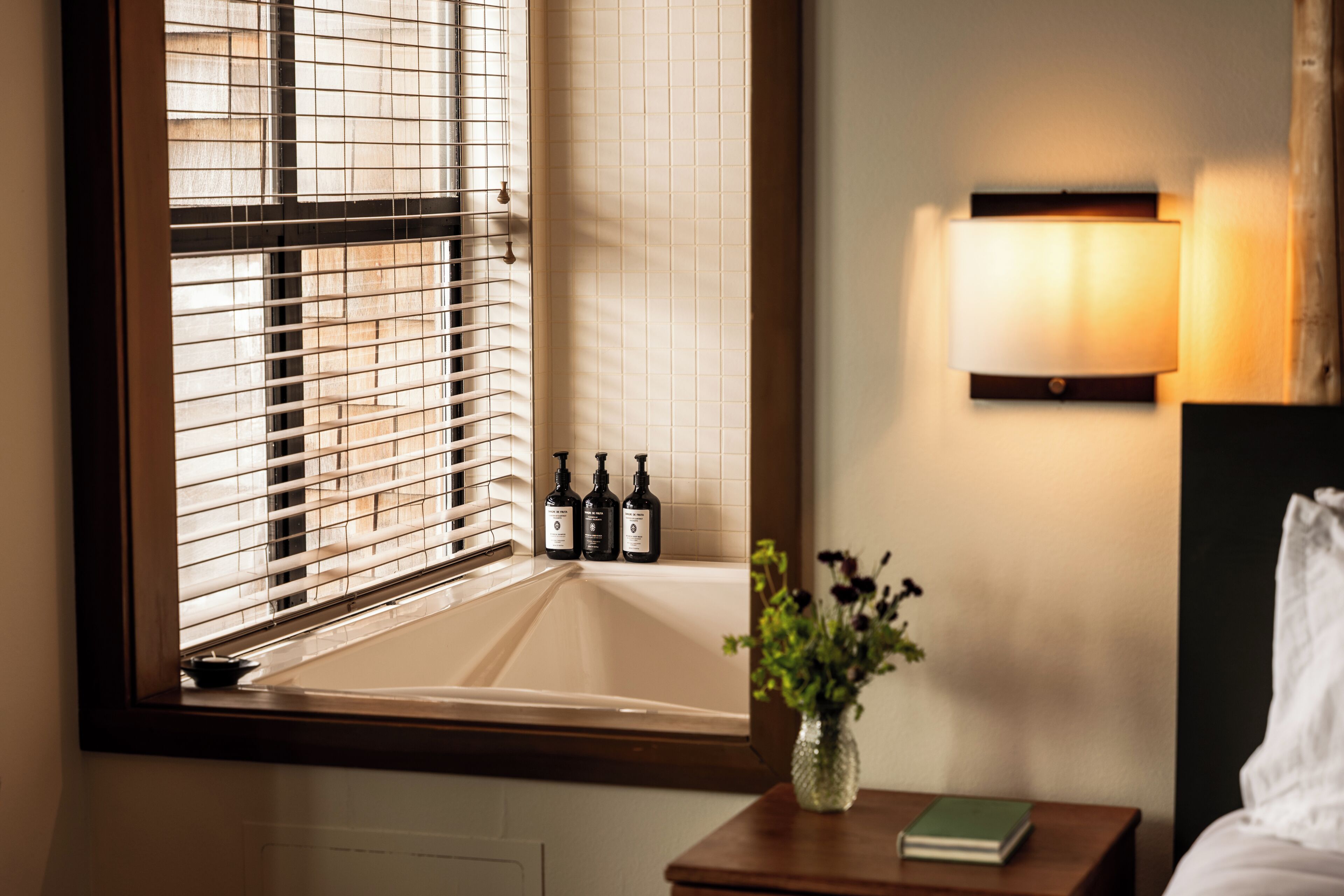 A bathroom with a bathtub, a window with blinds, a wall-mounted light fixture, and a small table with a vase and a book.