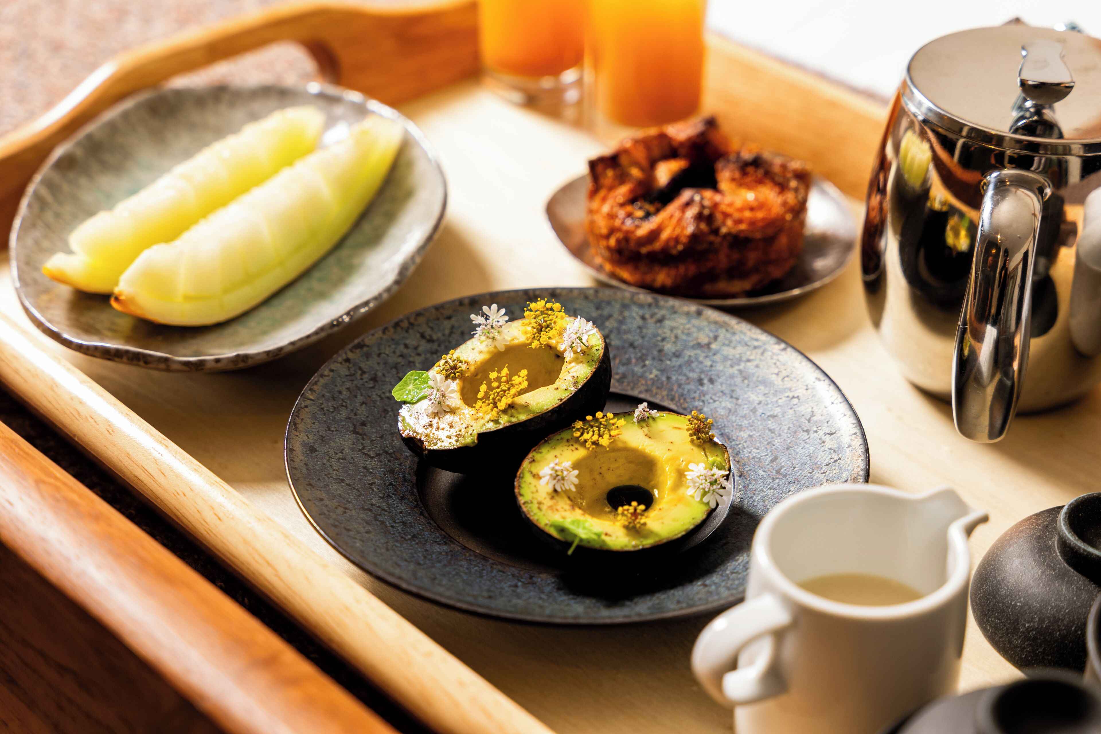 A breakfast tray with a plate of avocado halves, a bowl of fruit, a pastry, a glass of orange juice, and a coffee pot.