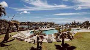 Zona de piscina con sillas de descanso y palmeras, rodeada por una valla y vista al océano.