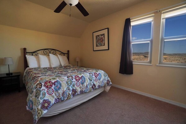 A bedroom with a bed, a nightstand, a ceiling fan, a window with a view of the desert, and a framed picture on the wall.
