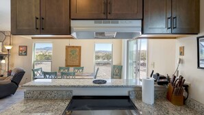 A kitchen with granite countertops, a built-in stove, and wooden cabinets. There is a dining area with chairs and a table, and a window with a view of the desert. - Lajitas Casa Grande: Spacious Four Bedroom Home (Terilingua)