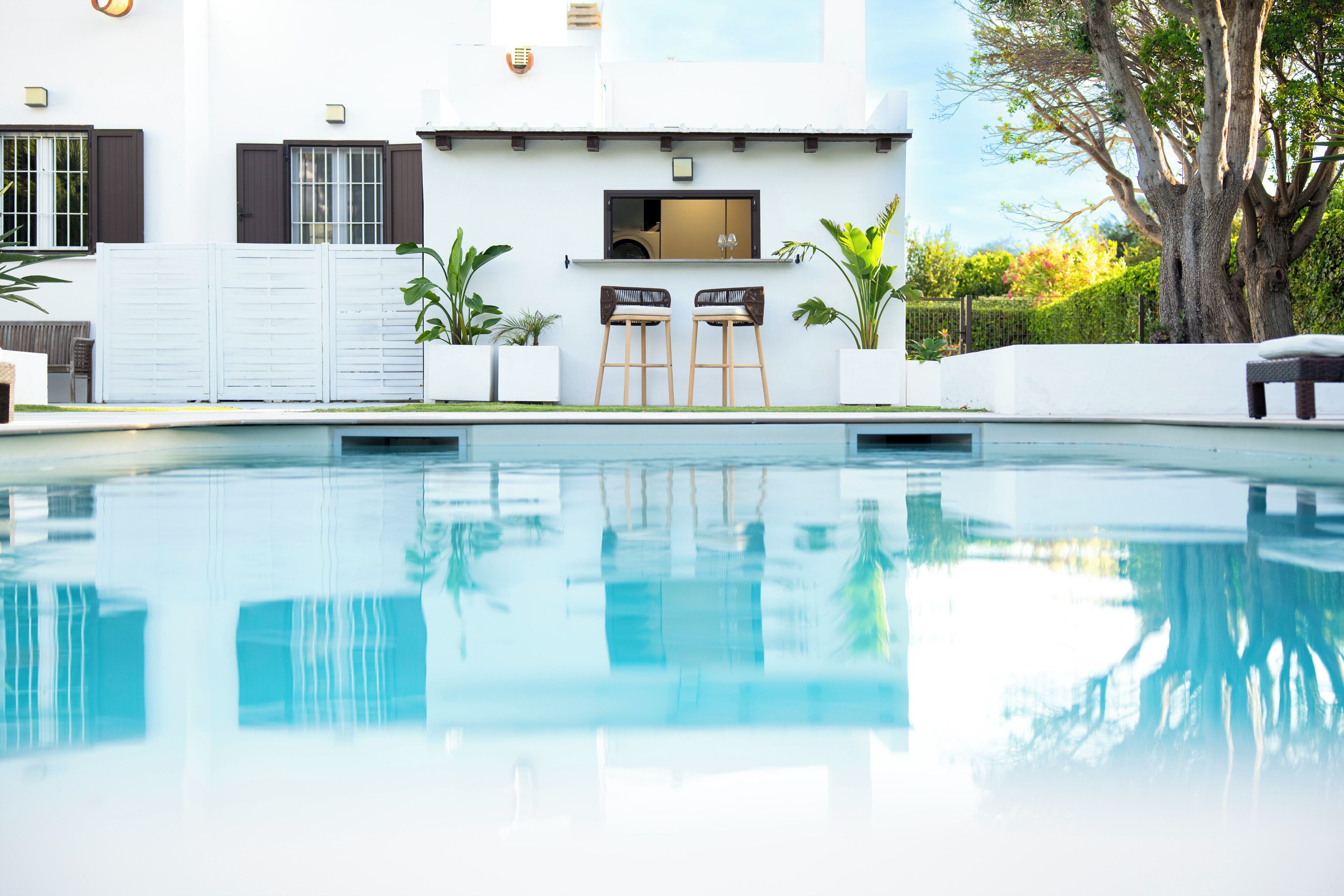 Une piscine avec un espace bar, un bâtiment blanc doté de volets et un grand arbre.
