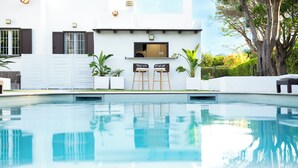 Una piscina con área de bar, un edificio blanco con persianas y un árbol grande.