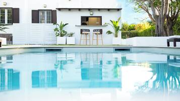 Une piscine avec un espace bar, un bâtiment blanc doté de volets et un grand arbre.