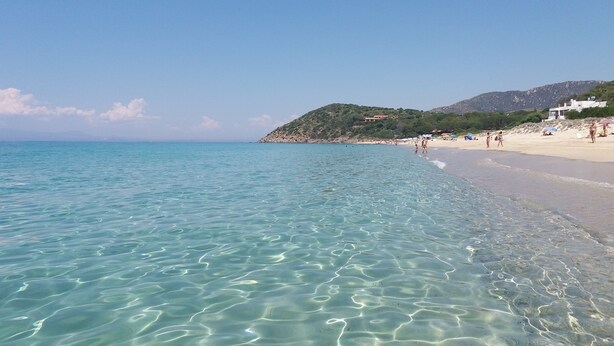 Una playa con aguas turquesas cristalinas, una orilla arenosa y un paisaje montañoso al fondo.