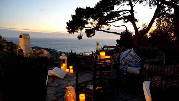 Ein Sitzbereich im Freien mit Korbmöbeln, Kerzen und Blick auf das Meer bei Sonnenuntergang.