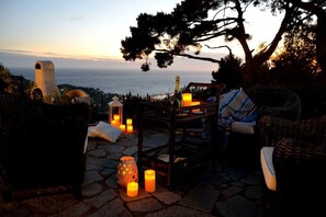 Un área de estar al aire libre con mobiliario de mimbre, velas y vista al mar al atardecer.