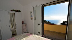 Ein Schlafzimmer mit Bett, Kommode, Kleiderschrank und einer Glastür, die auf einen Balkon mit Meerblick führt.
