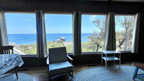 Una habitación con un ventanal grande que ofrece vistas al océano y un edificio lejano. Hay una silla y una mesita con mantel estampado.