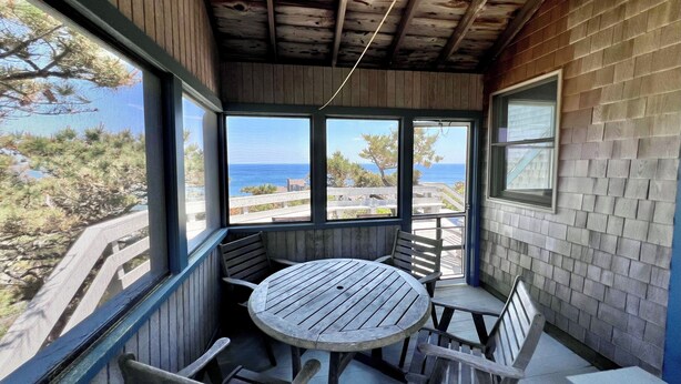 Una terraza de madera con una mesa redonda y bancos, con vistas a una zona costera con árboles y el mar.