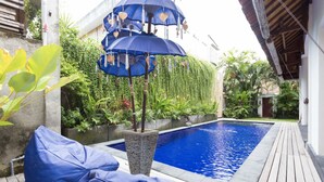 Una piscina con cubierta de madera, rodeada de exuberante vegetación y una estructura azul decorativa tipo sombrilla.