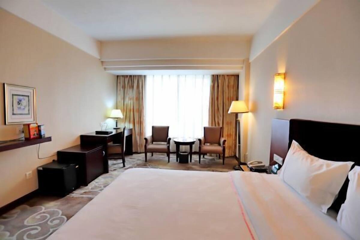 A hotel room with a large bed, a desk with a lamp, two chairs, a small table, and a window with curtains.