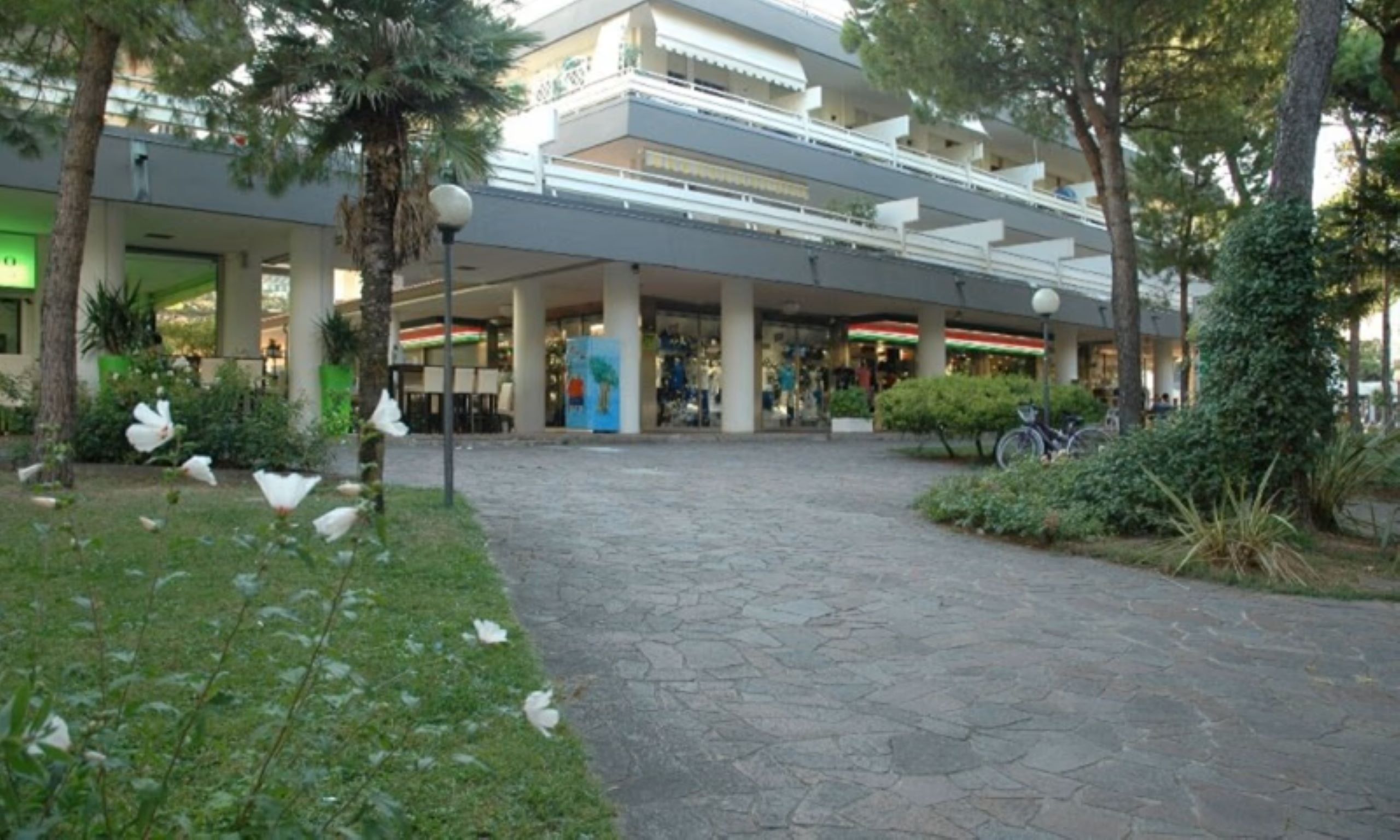 A multi-level shopping mall with a stone-paved walkway, surrounded by greenery and palm trees.