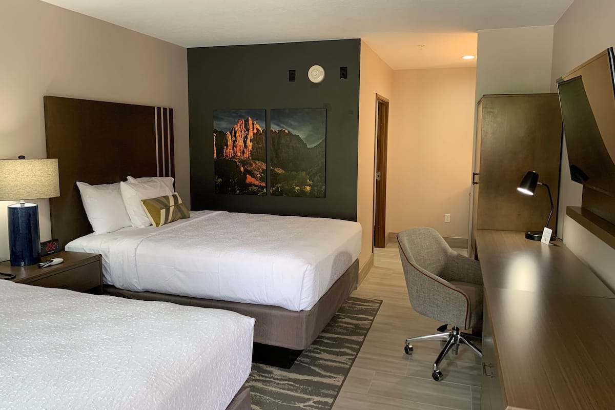 A hotel room with two beds, a desk, a chair, a lamp, and a painting on the wall.