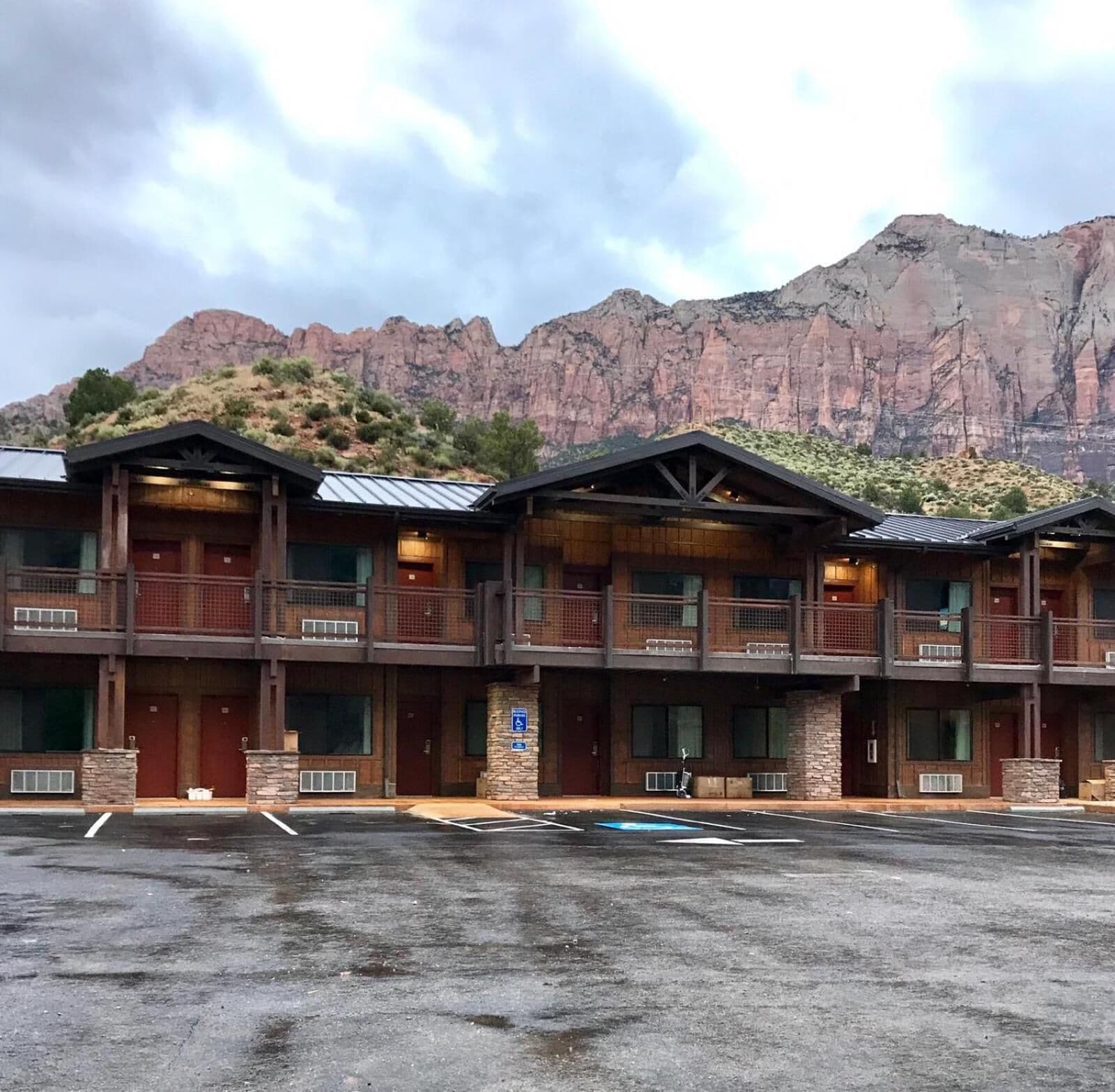 A multi-story hotel with a mountain backdrop. It features a central entrance with a covered walkway, multiple balconies, and a parking lot in front.