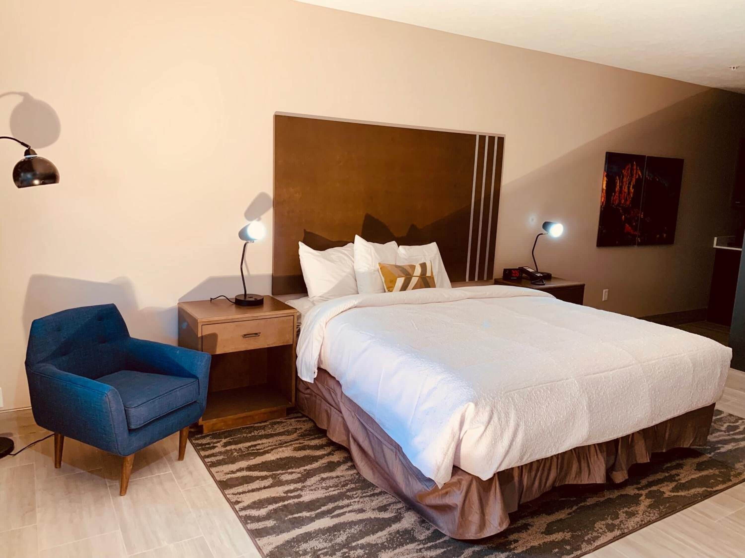 Ein Hotelzimmer mit einem großen Bett, einem Nachttisch mit Lampe, einem blauen Sessel und einem Bild an der Wand.