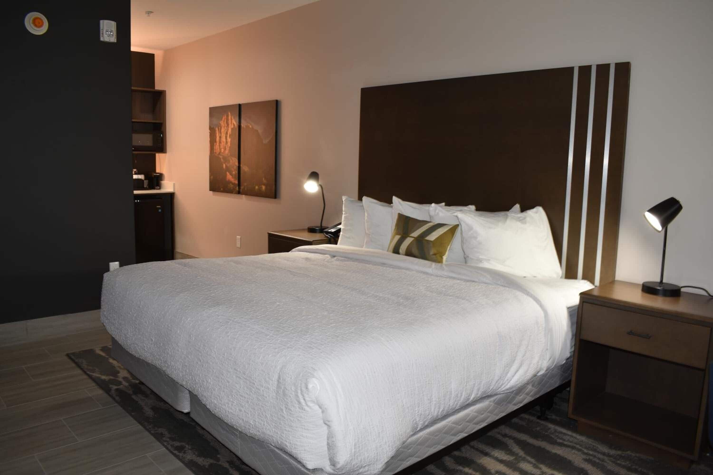 A neatly made bed with white bedding, a large headboard, bedside lamps, and a painting on the wall.