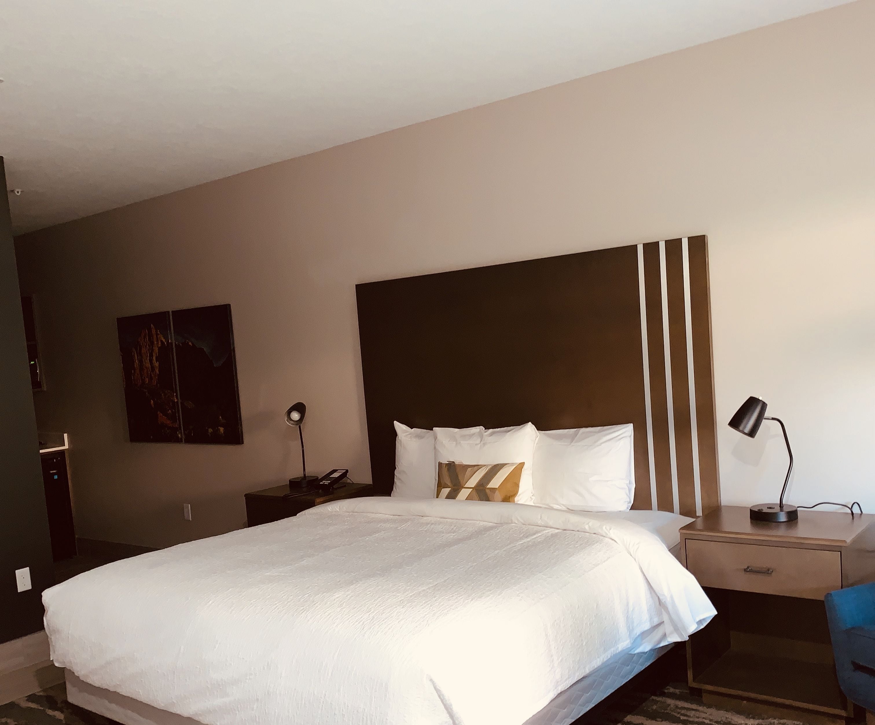 A hotel room with a large bed, a desk with a chair, a nightstand, and a wall-mounted lamp.