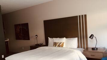 A hotel room with a large bed, a desk with a chair, a nightstand, and a wall-mounted lamp.