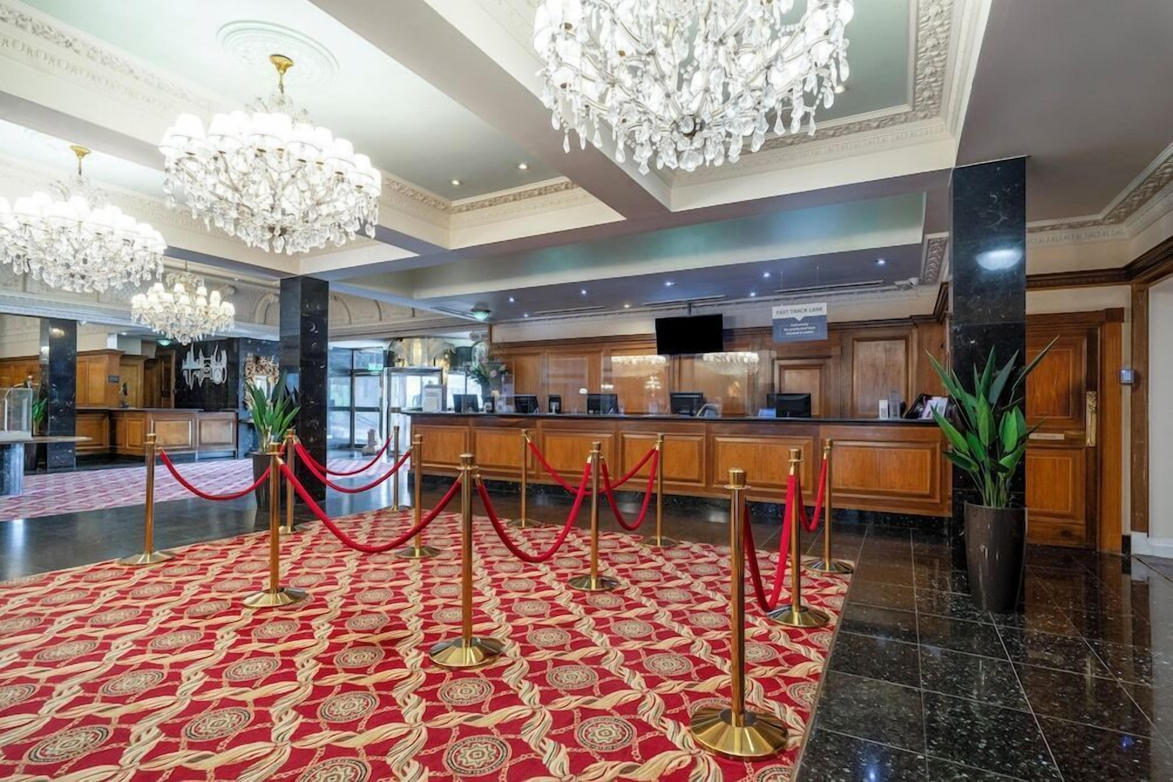 A hotel lobby with a red carpet, chandeliers, a reception desk, and a television screen.