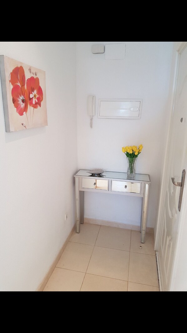 A hallway with a white door, a desk with a vase of yellow flowers, and a painting of red flowers on the wall. - FRONT APARTMENT FIRST LINE OF THE SEA (Orpesa)