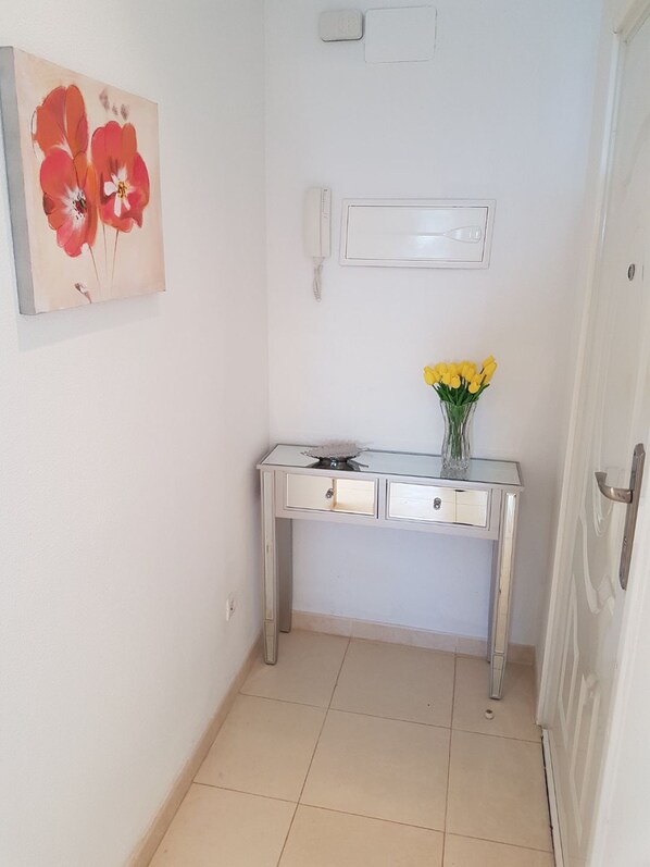 Un couloir avec une porte blanche, un bureau sur lequel est posé un vase rempli de fleurs jaunes, et un tableau représentant des fleurs rouges accroché au mur.