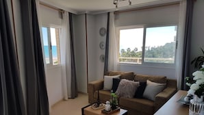 A living room with a sofa, a coffee table, and a view of the sea through a window.