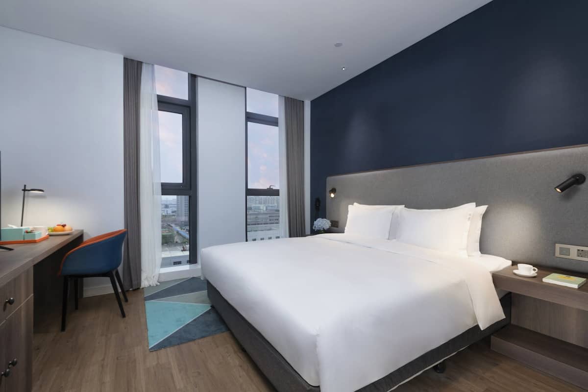 Ett modernt hotellrum med en stor säng, ett skrivbord med en stol, ett nattduksbord och utsikt över staden genom stora fönster.